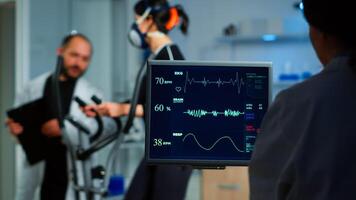 Medical researcher examining EKG image showing on monitor while patinet with mask running on cross trainer testing heart rate using electrodes.Doctor monitoring physical endurance in science sport lab photo