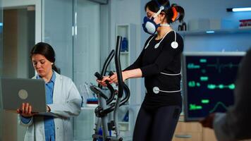 Medical researcher using laptop while measuring sportsman endurance using body sensors, electrodes and mask measuring cardiac rhythm. Patient running on cross trainer in sports science lab photo