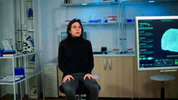 Stressed patient sitting on chair in neurological laboratory waiting for medical researcher investigating brain functions using high tech and neurology tools. Nervous system scan showing on monitor photo