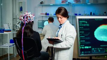 Back view of woman patient wearing performant brainwave scanning headset sitting in medical neurological laboratory while researcher adjusting it, examining nervous system typing on tablet. photo
