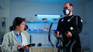 Man athlete running on cross trainer with electrodes attached to his body and mask. Physician using tablet computer and controls EKG data showing on laboratory monitors, discussing with patient photo
