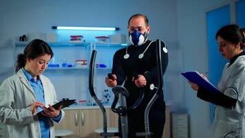 Athlete with mask and electrodes attached on body training with cross trainer while sport scientist interacs with tablet and supervises EKG status. Team of researchers using high-tech equipment photo