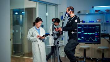 Sportsman with mask running on cross trainer in sports science laboratory measuring VO2 max, heart rate, psychological resistance and muscular endurance. Monitor showing EKG reading of sportsman photo