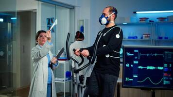 Medical researcher examining evolution of athlete's health condition, looking at x-ray while man with mask running on cross trainer monitoring his endurance. Monitor shows EKG reading of sportsman photo