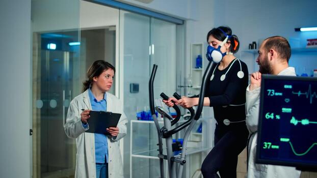 Sportswoman running with mask on cross trainer with electrodes attached to body. Physician using tablet computer and controls EKG data showing on laboratory monitors, discussing with patient photo