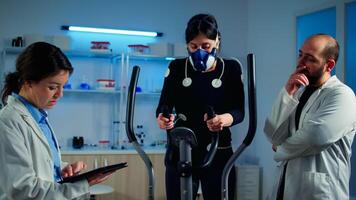 Woman athlete with mask doing fitness exercises in science sport lab with electrodes attached while scientist holding tablet supervising whole process. Physician using notepad controling EKG data photo