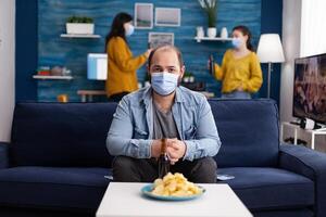 Young relaxed man wearing face mask looking at camera as safety prevention sitting on couch while speding time with friends during global pandemic with coronavirus. Group of young people having fun. photo