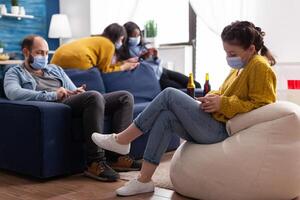 Group of mixed race friends browsing on smartphone keeping social distancing wearing face mask to prevent spread of coronavirus during global pandemic in living room with beer bottle. Conceptual image photo