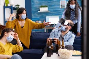 Group of multi ethnic friends having fun playing games experiencing virtual reality using vr headset keeping social distancing in home living room due to social pandemic. photo