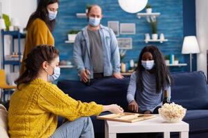 Group of cheerful multiethnic friends playing board games backgammon board games keeping social distancing wearing face mask to prevent infection with covid19 during global pandemic drinking beer. Conceptual image. photo