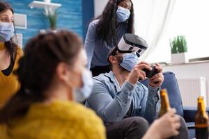 Excited man spending time with friends experiencing vritual reality playing games with vr headset wearing face mask to prevent coronavirus spread in time of social pandemic. photo
