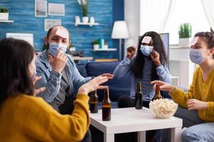 Cheerful friends with sticky notes on foreheads playing name game with beer popcron keeping social distacing with face mask to prevent spread of covid19 in home living room. Conceputal image. photo