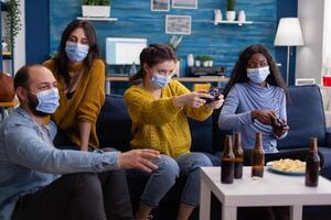 Concentrated multiethnic friends trying to win at games during social pandemic wearing face mask keeping distancing not to spread corona sitting on couch in living room. photo