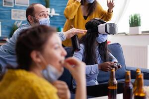 African woman experiencing virtual reality playing games with vr headset and joystick in home living room while friends are cheering up for her keeping social distancing because of global pandemic. photo