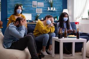 Group of mixed race friends losing while playing games in home living room with joystick wearing face mask keeping social distancing in time of corona outbreak. Enoying beer and chips. photo