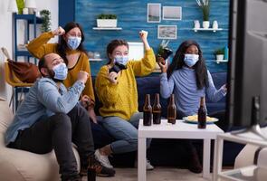Excited group of multiethnic friend celebrating victory while playing games in home living room keeping social distancing wearing face mask not to spread corona in time of global pandemic. photo