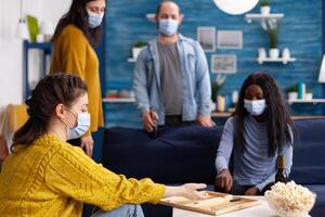 Cheerful happy woman using dice playing backgammon with multiethnic friends wearing face mask as prevention for covid 19 spread during global pandemic sitting on couch drinking beer and eating popcorn. photo