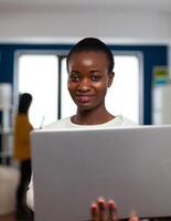 Portrait of african woman looking at camera smiling standing in start up multimedia design agency holding laptop, typing on it. Black maker editing creative montage using production software photo