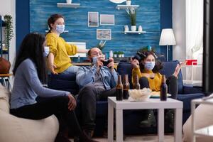 Excited group of multi ethnic friends scraming celebrating victory during gaming competition wearing face mask keeping social distancing to prevent coronavirus spread in time of global pandemic. photo
