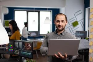 creative editor worker standing in front of camera smiling working in digital agency office holding laptop. Man videographer works in multimedia post production studio editing content photo