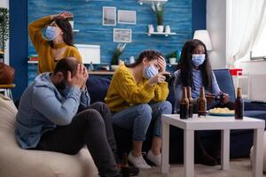 Disappointed diverse group of people playing games respecting social distancing because of orona outbreak wearing face mask not to spread the virus and get sick. Beer bottle and chips. photo