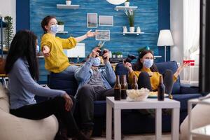 Group of multiethnic friends having a gaming competition in apartment home living room using wireless joystick sitting on sofa keeping social distancing wearing face mask in the course of coronavirus global outbreak. photo