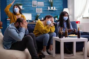 Sad multi ethnic group of friends after lose at gaming competiton with face covers with mask during covid outbreak keepin social distancing in home living room. Enjoying beer and chips. Conceptual image. photo