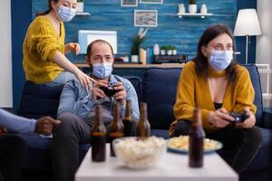Diverse friends trying to win playing games using joystick havin fun wearing face mask to prevent spreading of coronavirus in time of global outbreak. Gaming competition, beer and popcorn. photo