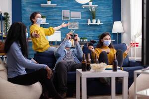 Happy cheerful woman screaming after wining gaming competiton with multiethnic friends in home living room with face mask preventing coronavirus spreading in time of global pandemic. photo