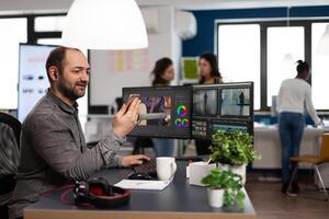 Multimedia creator artist talking on videocall with client holding smartphone editing footage sitting in creativity production agency. Videographer using post production software photo