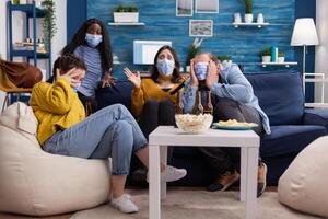 Group of multi ethnic friends watching a horror movie on tv being afraid wearing face mask to prevent infection with covid 19 during global pandemic having fun sitting on couch and photo