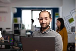 creative editor worker standing in front of camera smiling working in digital agency office holding laptop. Man videographer works in multimedia post production studio editing content photo