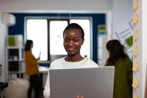 Portrait of african woman looking at camera smiling standing in start up multimedia design agency holding laptop, typing on it. Black maker editing creative montage using production software photo