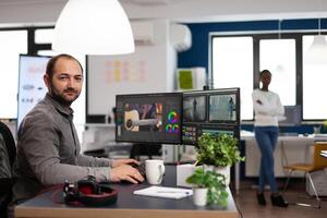 Creative videographer artist man looking at camera smiling working in creativity multimedia startup office using editing post production software on professional pc. Processing film montage. photo