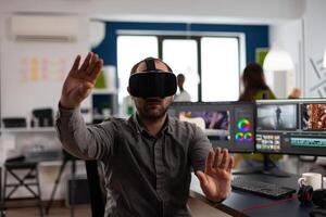 Designer creator wearing VR headphones gesturing while editing montage project in post production software sitting in studio office. Man videographer processing movie in digital multimedia company photo