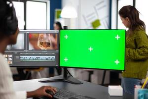 African designer editor with headset editing footage using pc with greenscreen, chroma key isolated display sitting in agenc company. Videographer in digital multimedia company processing movie photo