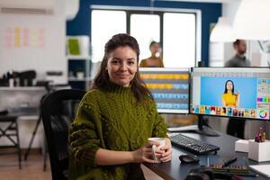 Woman retoucher looking at camera smiling sitting in creative design media agency retouching client photos on PC with two displays. Artist graphic editor working in start up office with digital assets