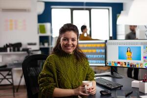 Woman retoucher looking at camera smiling sitting in creative design media agency retouching client photos on PC with two displays. Artist graphic editor working in start up office with digital assets