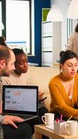 Businesswoman corporate leader talking to diverse team explaining project plan reviewing paperwork sitting on couch. Multiethnic coworkers taking notes on laptop and tablet analysing financial reports photo