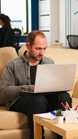 Business man typing on laptop sitting on couch in start up office while diverse team working in background analysing statistics data. Multiethnic coworkers talking about startup financial company photo