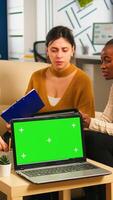 Diverse women sitting at table in start up office discussing strategy with green screen laptop in front of camera. Lady explaining to multiethnic team new project, chroma key display. mock up monitor photo