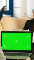 Over the sholder of manager sitting on couch talking with colleague holding laptop with green screen while diverse team working on background. Multiethnic people planning project on chroma key display photo