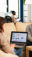Back view of young business woman sitting on couch in office using laptop computer while colleagues working on background. Multiethnic coworkers planning new financial project in modern company photo