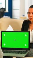 Business team sitting on couch analysing financial statistics, holding laptop with green screen while diverse team working on background. Multiethnic coworkers planning project on chroma key display photo