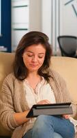 Employee taking break sitting on confortable couch holding tablet browsing, reading great news and smiling. Multiethnic creative coworkers planning new financial project working in open plan office photo