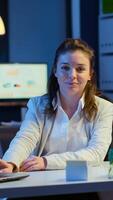 Business woman looking at camera smiling after typing on laptop sitting at desk in start-up company late at night. Focused employee using technology network wireless doing overtime for job reading. photo
