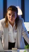 Business woman looking at camera smiling after typing on laptop standing near desk in start-up company late at night. Focused employee using technology network wireless doing overtime for job reading. photo