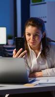 Business woman discussing during conference with partners using smartphone and headphone late at night working in start-up office. Employee with modern technology network wireless doing overtime photo