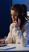 Woman employee speaking at phone while working at laptop late at night. Busy focused freelancer using modern technology network wireless doing overtime for job reading writing, searching taking break photo
