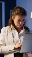 Focused businesswoman checking business reports standing in start up office late at night typing on laptop. Busy employee using technology network wireless doing overtime reading writing, searching photo